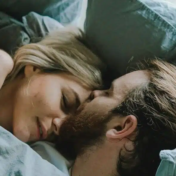 A bearded woman kissing a woman’s forehead while they lie on a bed.