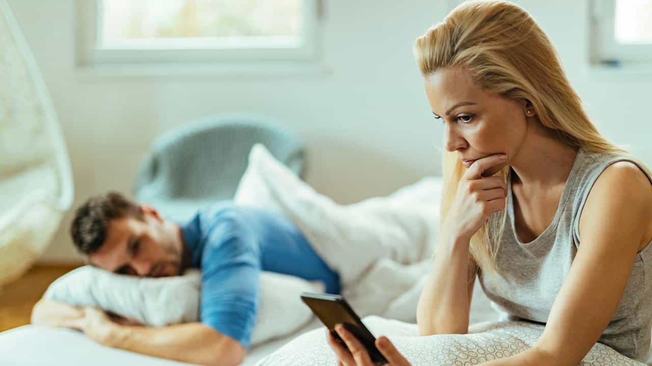A displeased woman reading texts on a phone while her boyfriend is sleeping.