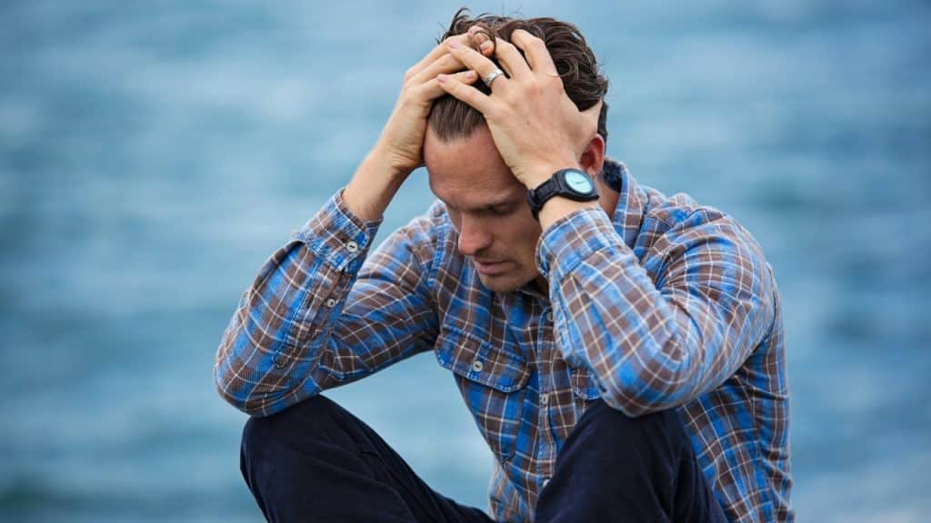 A man in a plaid shirt sits outdoors with both hands on his head.