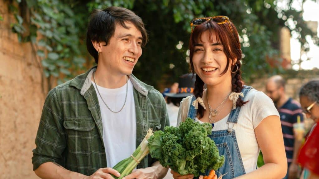 Two smiling people stand outdoors while holding bunches of kale and green onions.