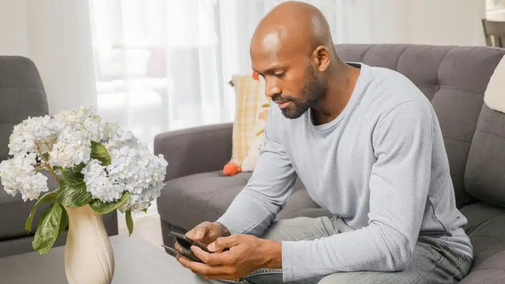 A bald man sits on a grey sofa looking down at his mobile phone screen.
