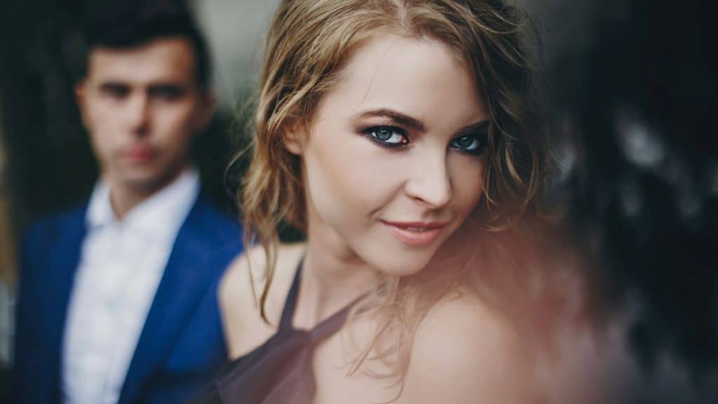 Close-up of a woman with blonde hair and smoky eye makeup; blurred man in background.