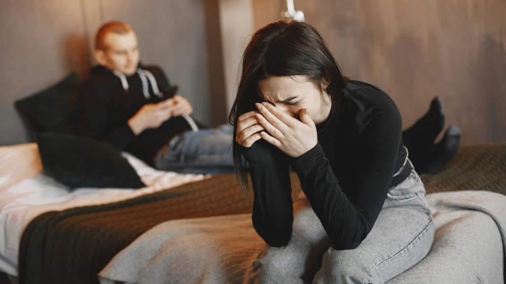 A woman cries with her face in her hands while a man sits in the background on his phone.