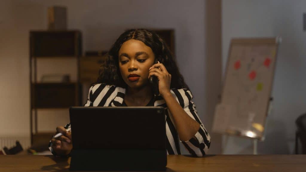A woman in a striped blazer talks on a phone while looking at a tablet.