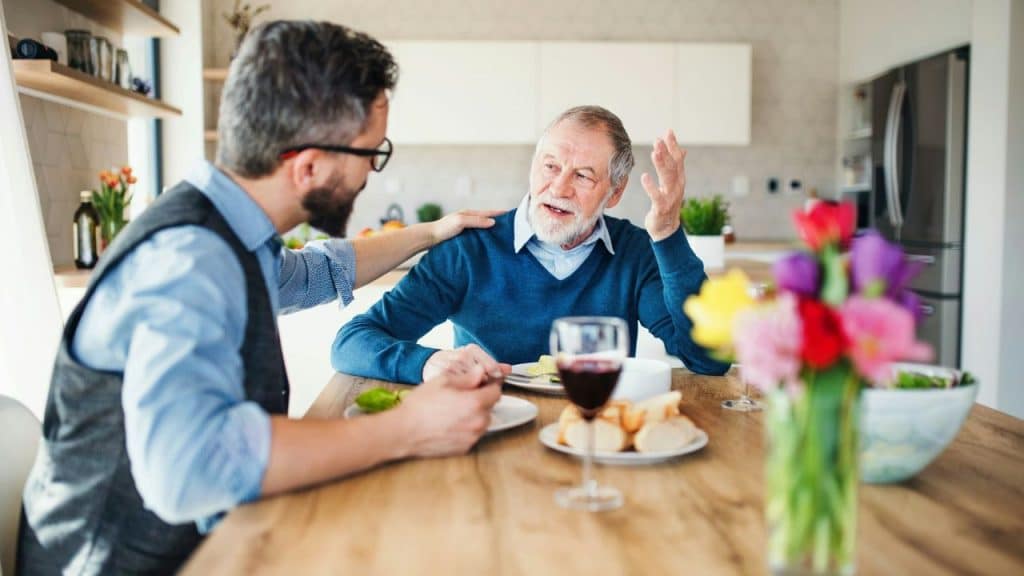 Two men sit at a dining table with food, one gesturing while the other listens.