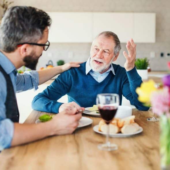 Two men sit at a dining table with food, one gesturing while the other listens.
