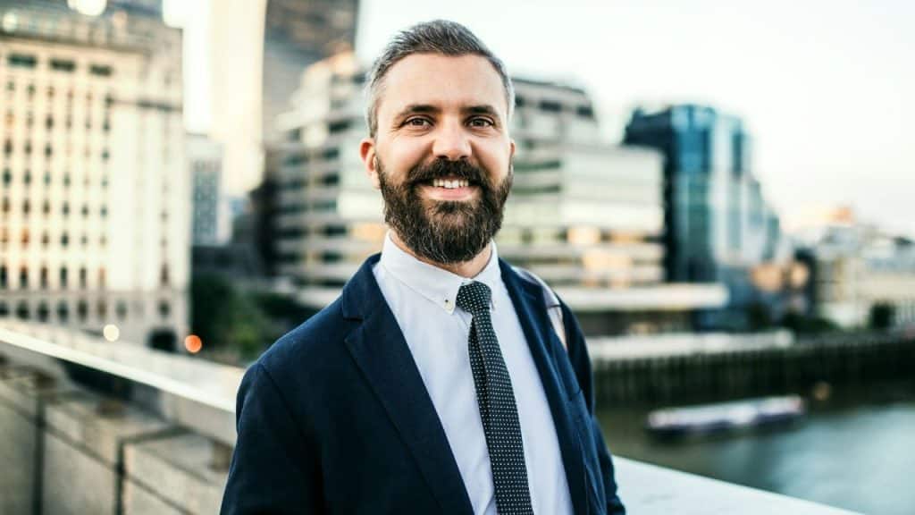 A smiling man with a beard wearing a suit and tie stands outdoors before city buildings.