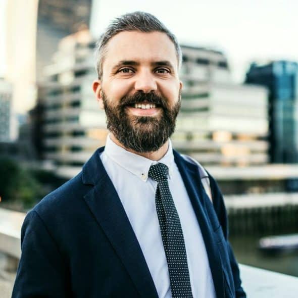 A smiling man with a beard wearing a suit and tie stands outdoors before city buildings.