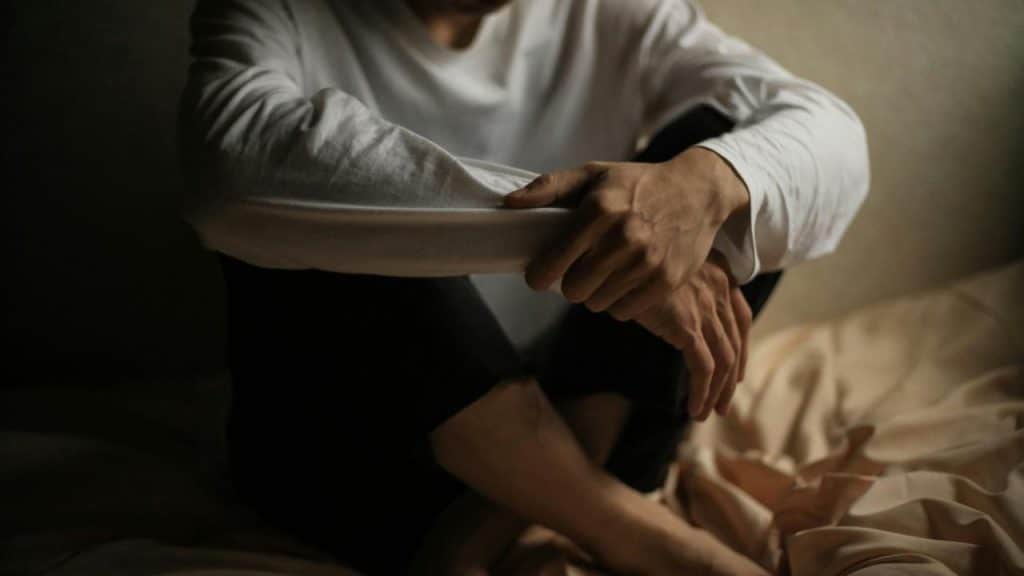 A person sitting with arms wrapped around their knees in a dimly lit room.