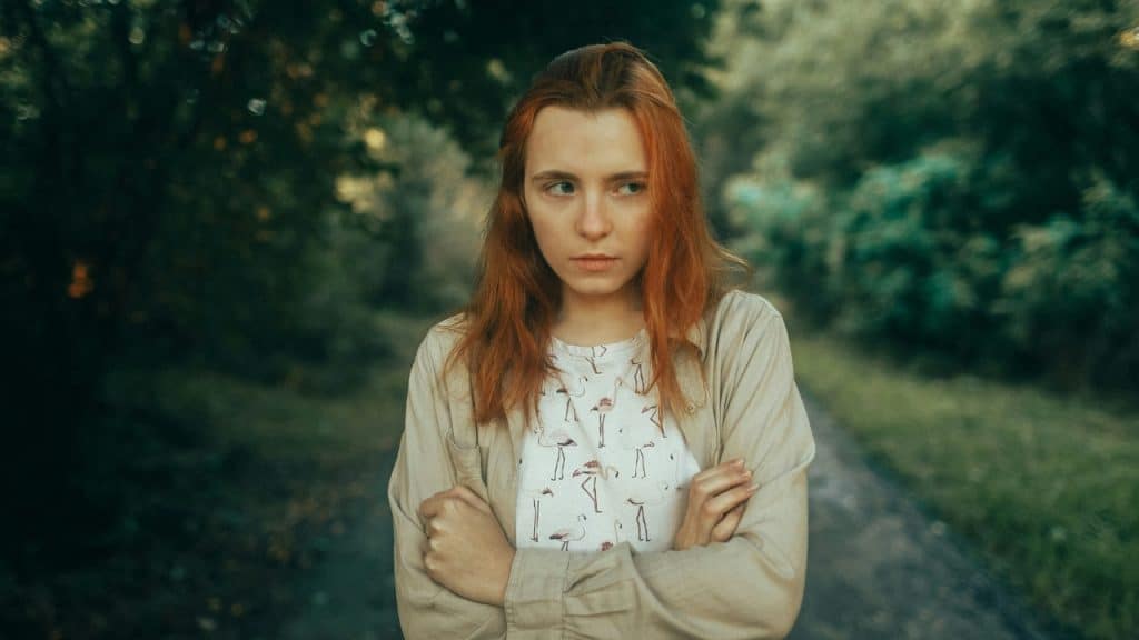 A young woman with red hair stands outside with arms crossed looking off to the side.