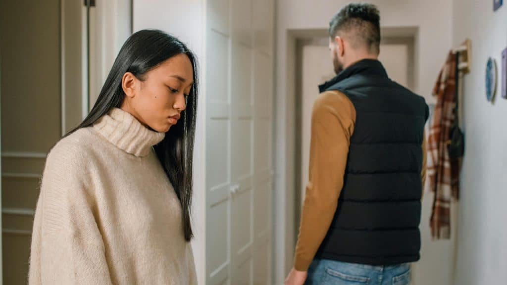 A woman looks down while a man in a black vest walks away down a hallway.