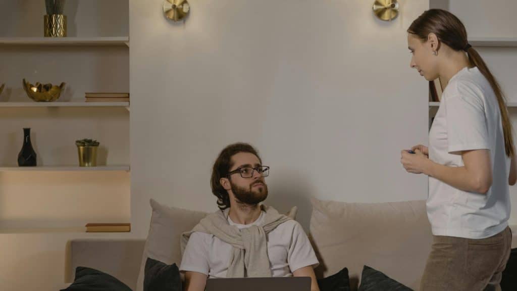 A man with a laptop looks up at a woman standing in a dimly lit room.