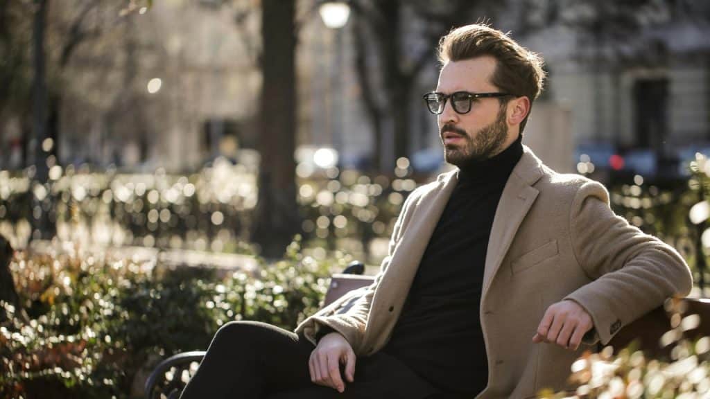 A man in a tan coat and black turtleneck sits on a park bench outdoors.