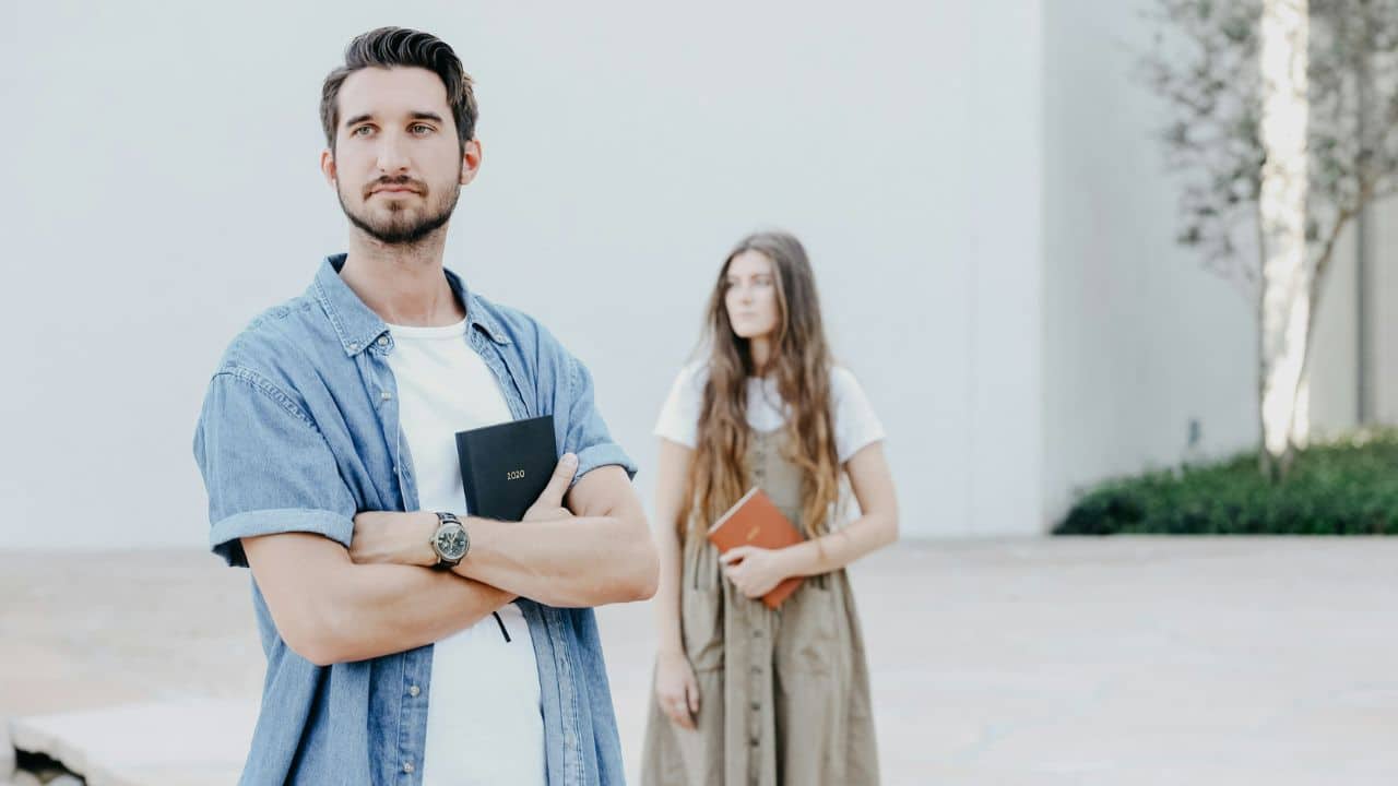 A man with crossed arms holds a small book while a woman stands in the background.
