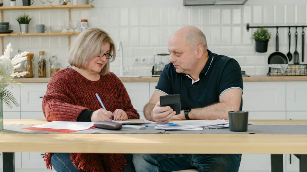 A man and woman sit at a kitchen table reviewing several papers and a calculator.
