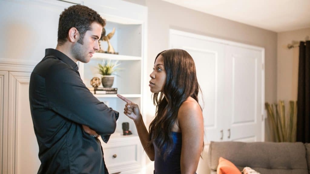 A woman points her finger toward a man standing with his arms crossed in a room.