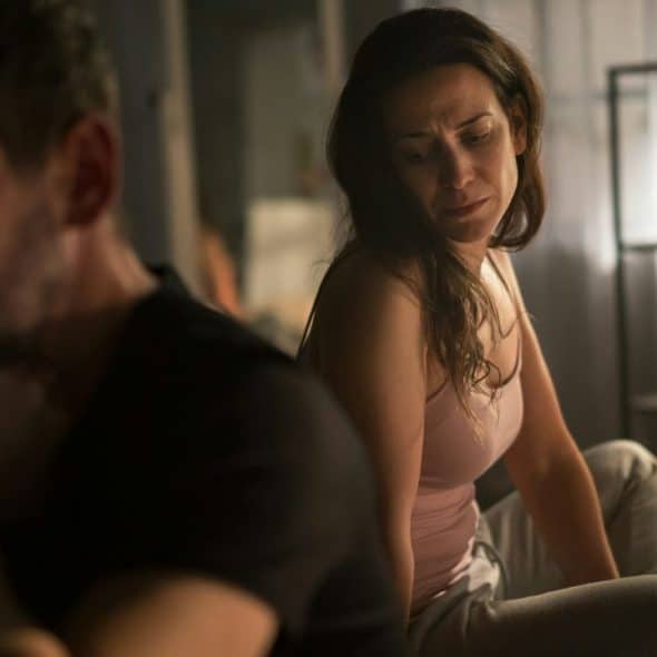A woman in a pink tank top sits on a bed looking at a man.