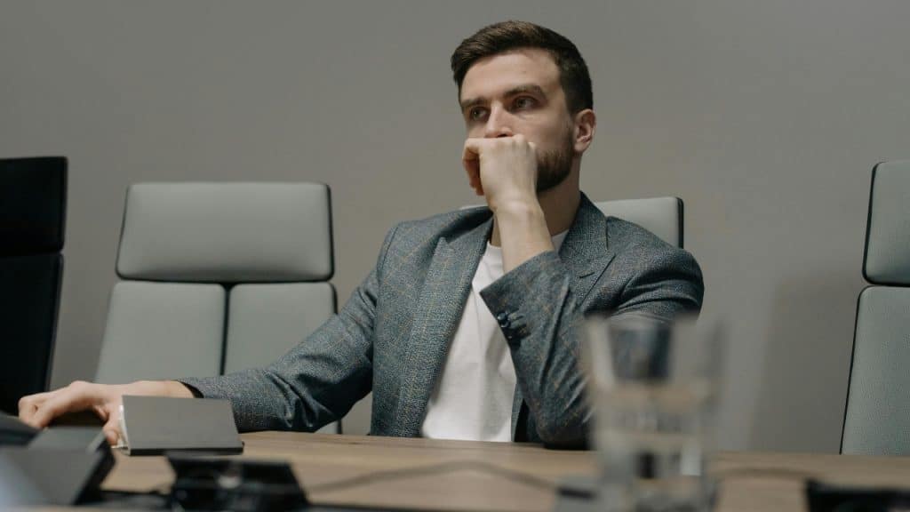 A man in a grey blazer sits at a wooden table with his hand on his chin.