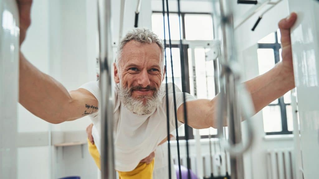 A smiling man with a grey beard performs an exercise on a gym cable machine.