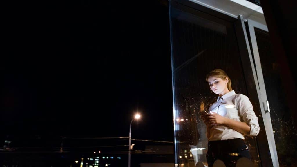 A woman standing by a window at night while looking at her phone.