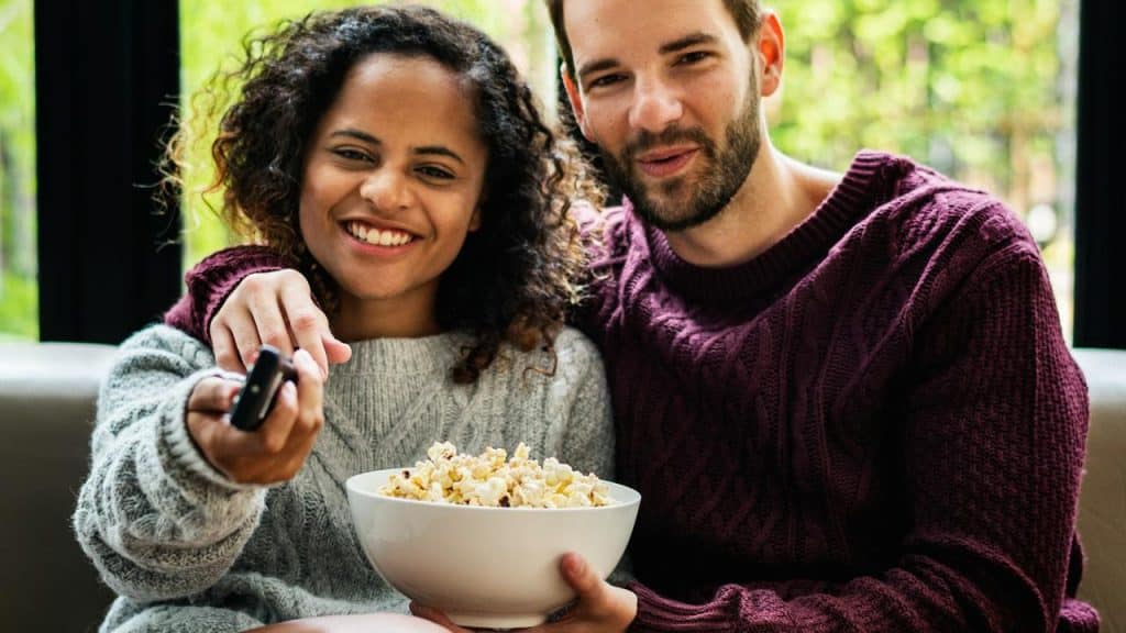 A couple smiling while watching TV and sharing popcorn.
