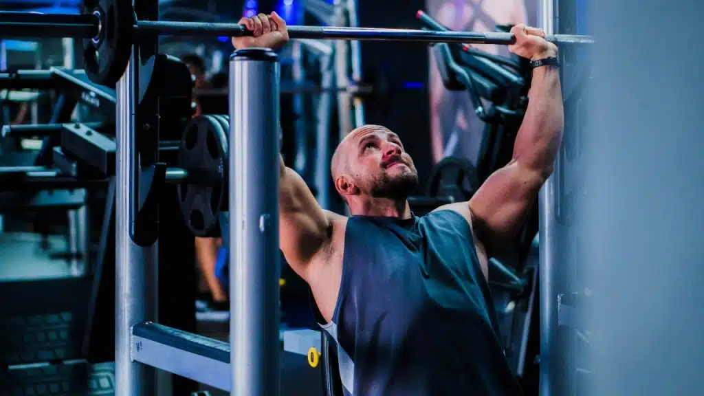 A man in a gym lifts a weighted barbell upward while looking toward the ceiling.