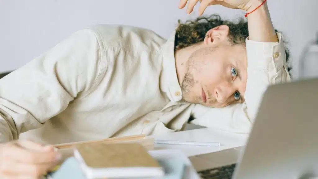 A tired man resting his head on a desk beside a laptop and notebooks.