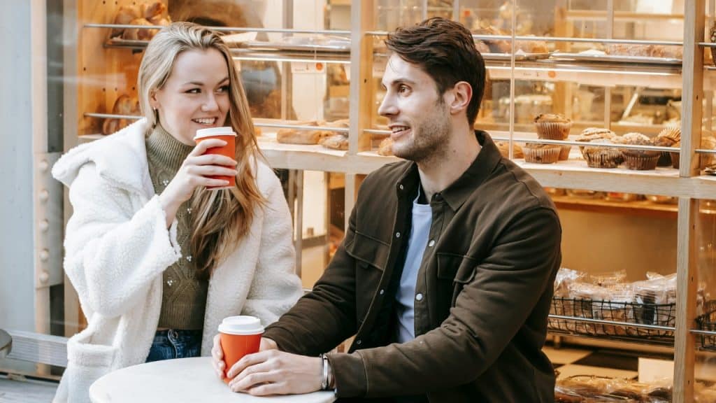 A man and woman sit at an outdoor table holding red coffee cups near a bakery.