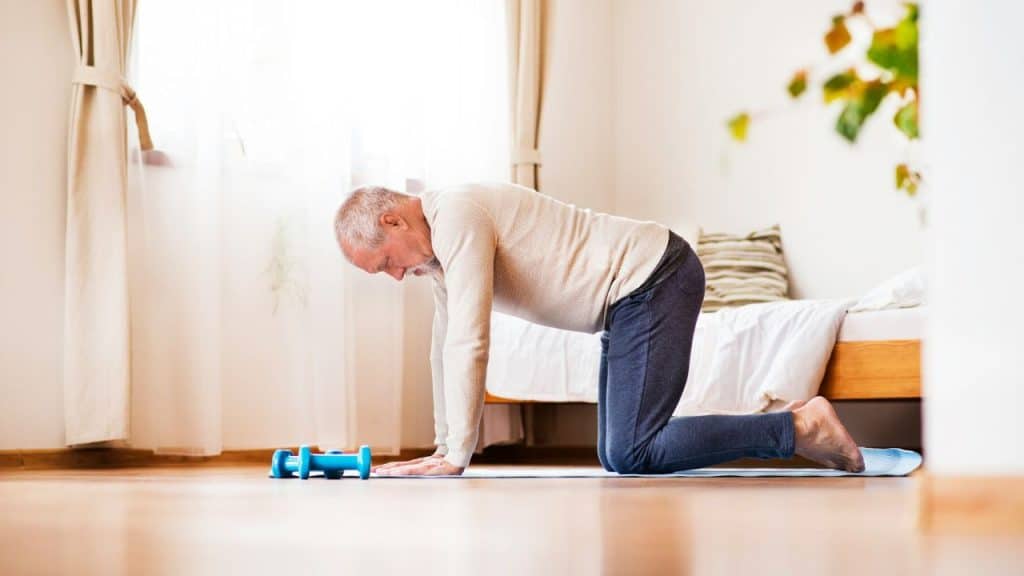A senior man on a yoga mat in a room performs a table-top stretch.