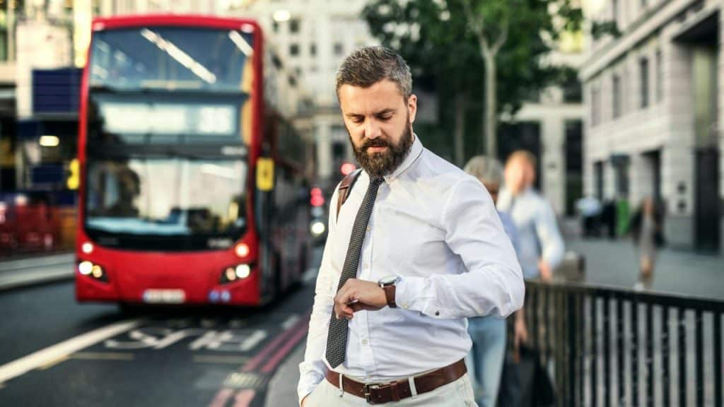 A bearded man in a white shirt and tie checks his watch on a city street.