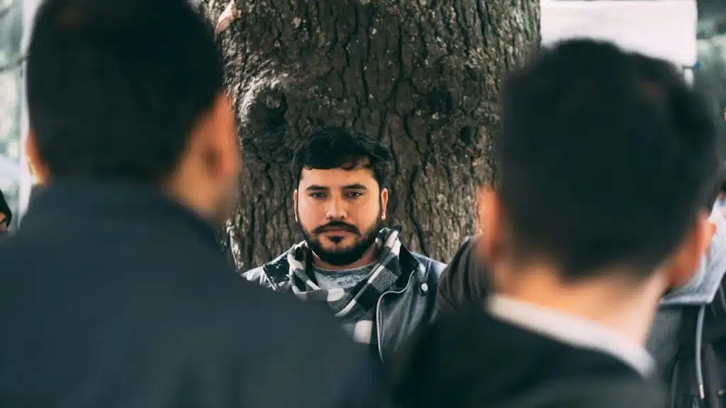 Man in gray-and-white-plaid scarf leaning on tree during daytime.