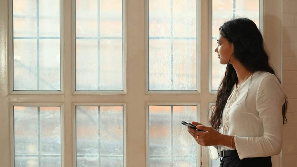 A beautiful woman is listening to music by smartphone in hand while standing next to living room windows.