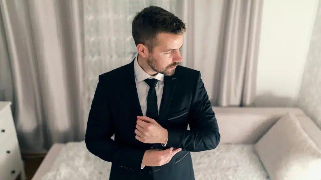 A man in a dark suit and tie adjusts his cufflink while looking to the side.