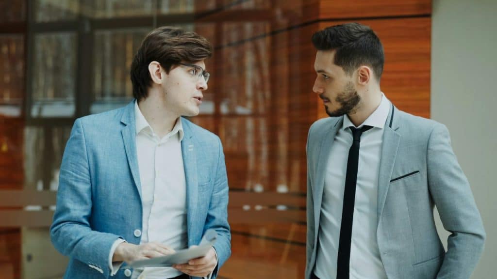 Two men in suits stand in an office hallway and hold some papers.
