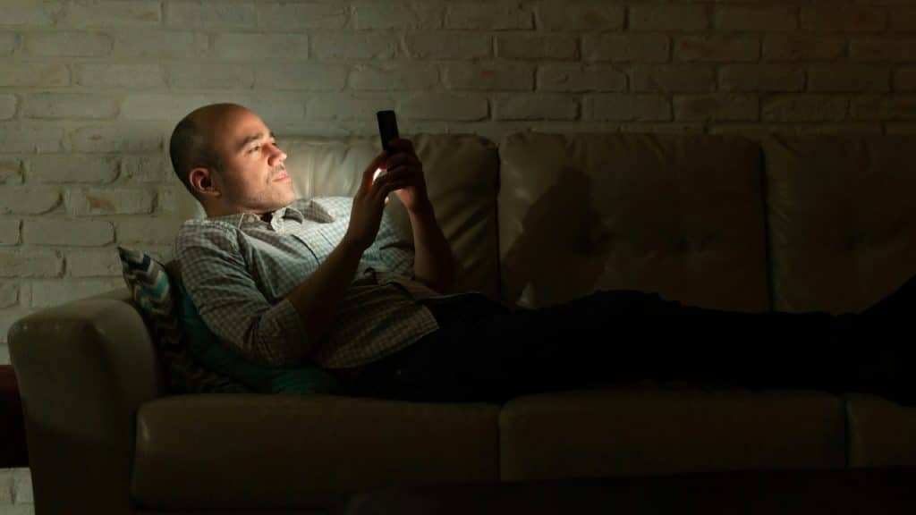 A man sits on a couch in the dark using his glowing smartphone.