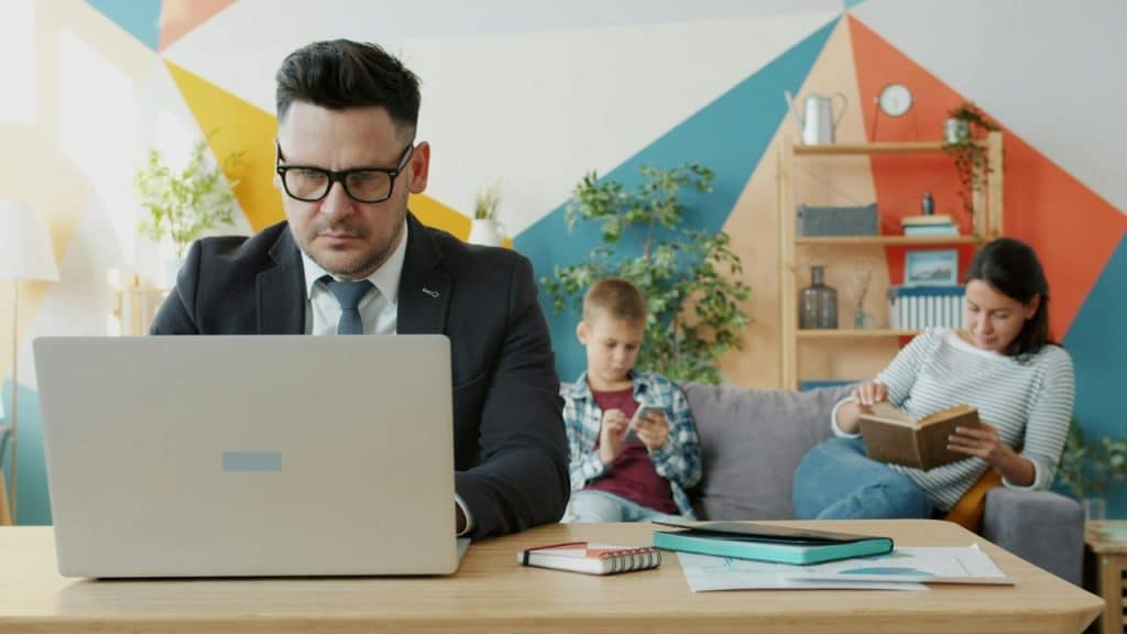 A man uses a laptop at a desk while a woman and child sit behind him.