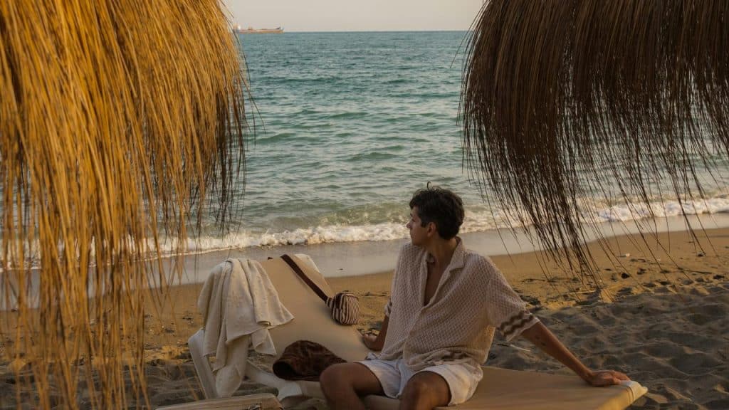 A person sitting on a beach lounger under thatched umbrellas, looking out at the sea.