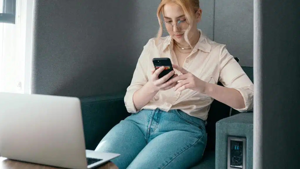 A woman using a smartphone while sitting beside a laptop.