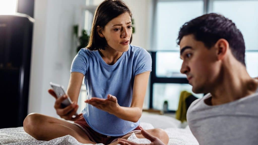 A woman sits on a bed holding a smartphone while gesturing toward a man nearby.