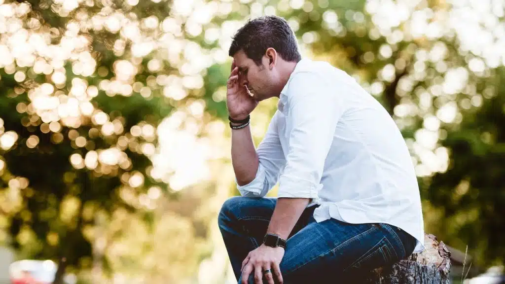 A man in a white shirt sits outdoors, resting his forehead on his hand sadly.