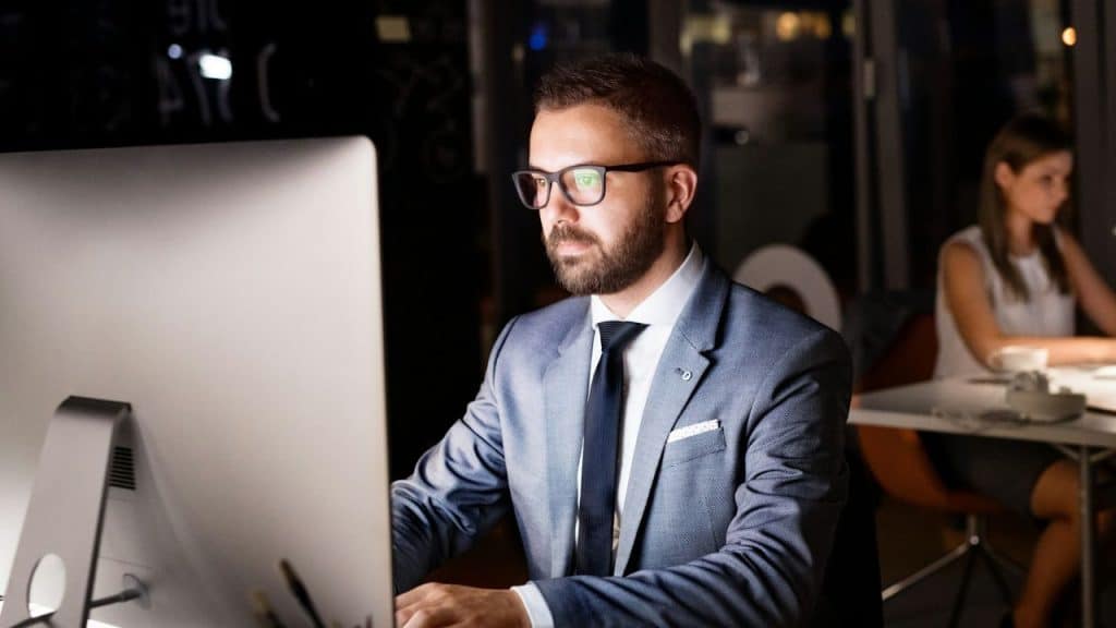 A man in a suit and glasses looks at a computer screen in a dark office.