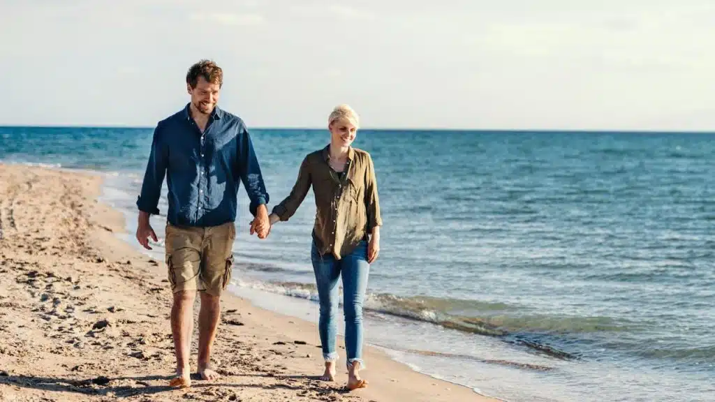 A man and woman hold hands while walking along a sandy beach near the ocean.