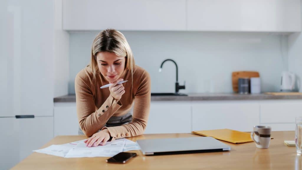 A woman in a brown sweater leans over a table reviewing various paper documents.