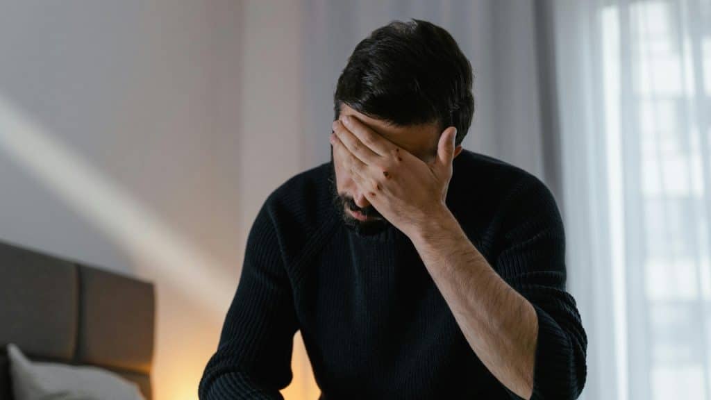 A man in a dark sweater sits indoors with his hand covering his forehead and eyes.