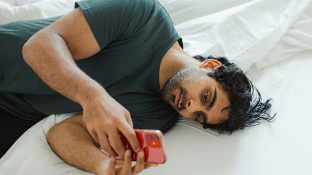 A man wearing a dark t-shirt lies on a white bed, holding a phone.