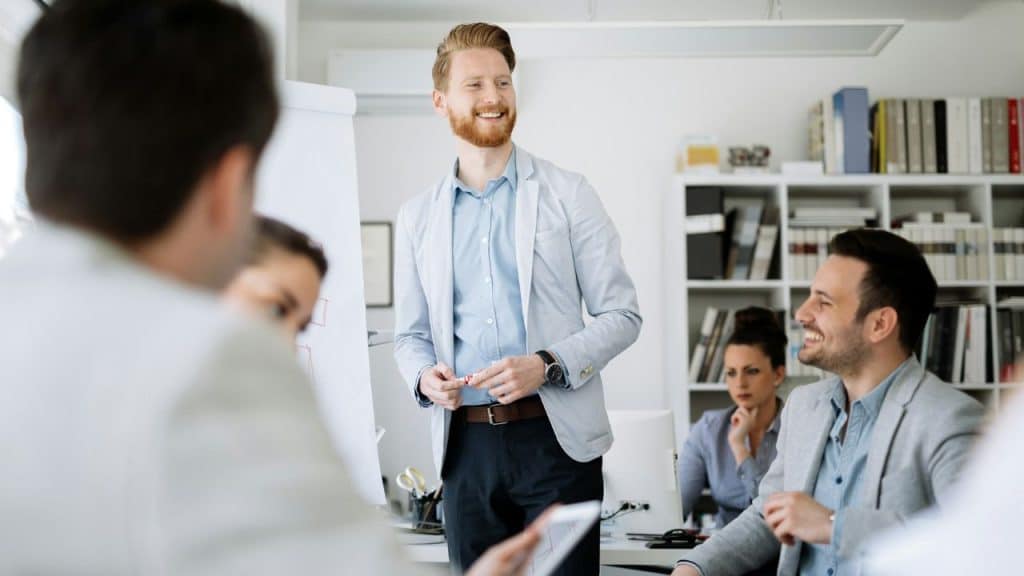 A man in a blazer stands before his colleagues while leading a professional meeting.