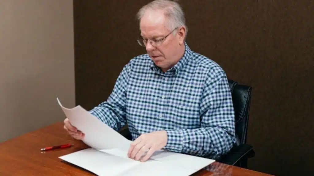 A man in a plaid shirt sits at a desk reading a document.