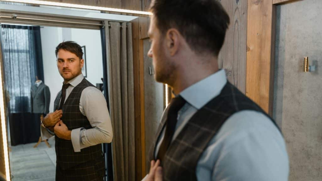 A man adjusts his suit vest while looking at his reflection in a mirror.