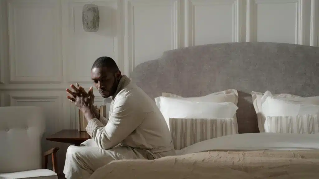 A man sitting on the edge of a bed with his hands clasped, looking thoughtful.