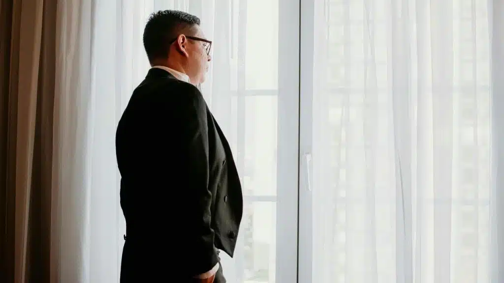 A man in a suit standing by a window and looking outside.